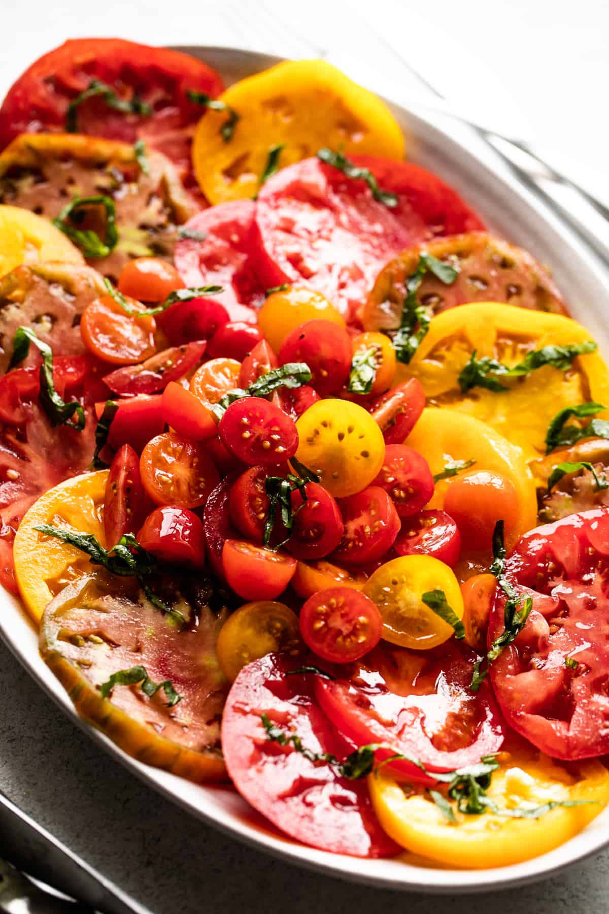 slices of heirloom tomatoes arranged on an oval platter, with halved cherry tomatoes placed in the center, and a garnish of basil over the entire salad.
