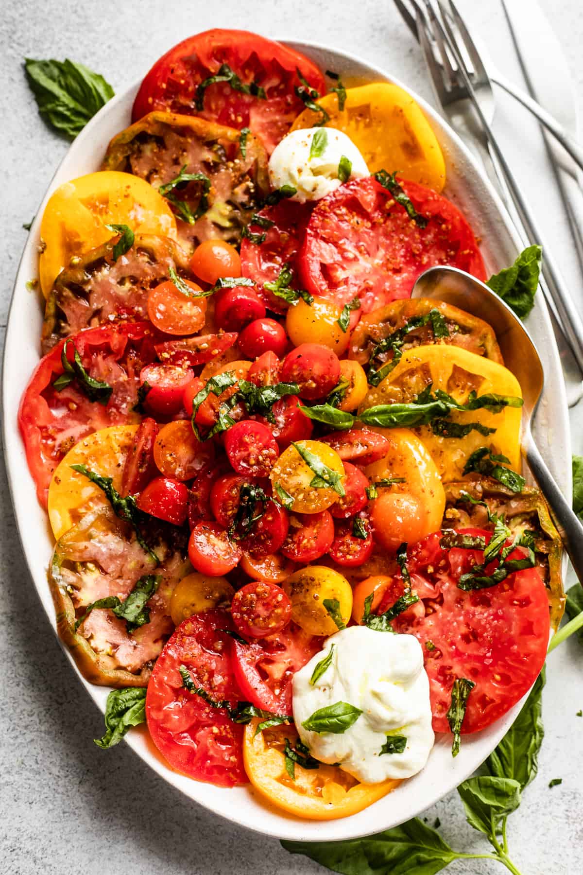 slices of heirloom tomatoes arranged on an oval platter, with halved cherry tomatoes placed in the center, burrata cheese at the top of the plate and the bottom, and a garnish of basil over the entire salad.