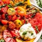 Up close shot of Tomato Burrata Salad: slices of heirloom tomatoes arranged on an oval platter, with halved cherry tomatoes placed in the center, burrata cheese at the top of the plate and the bottom, and a garnish of basil over the entire salad.