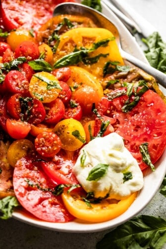 Up close shot of Tomato Burrata Salad: slices of heirloom tomatoes arranged on an oval platter, with halved cherry tomatoes placed in the center, burrata cheese at the top of the plate and the bottom, and a garnish of basil over the entire salad.