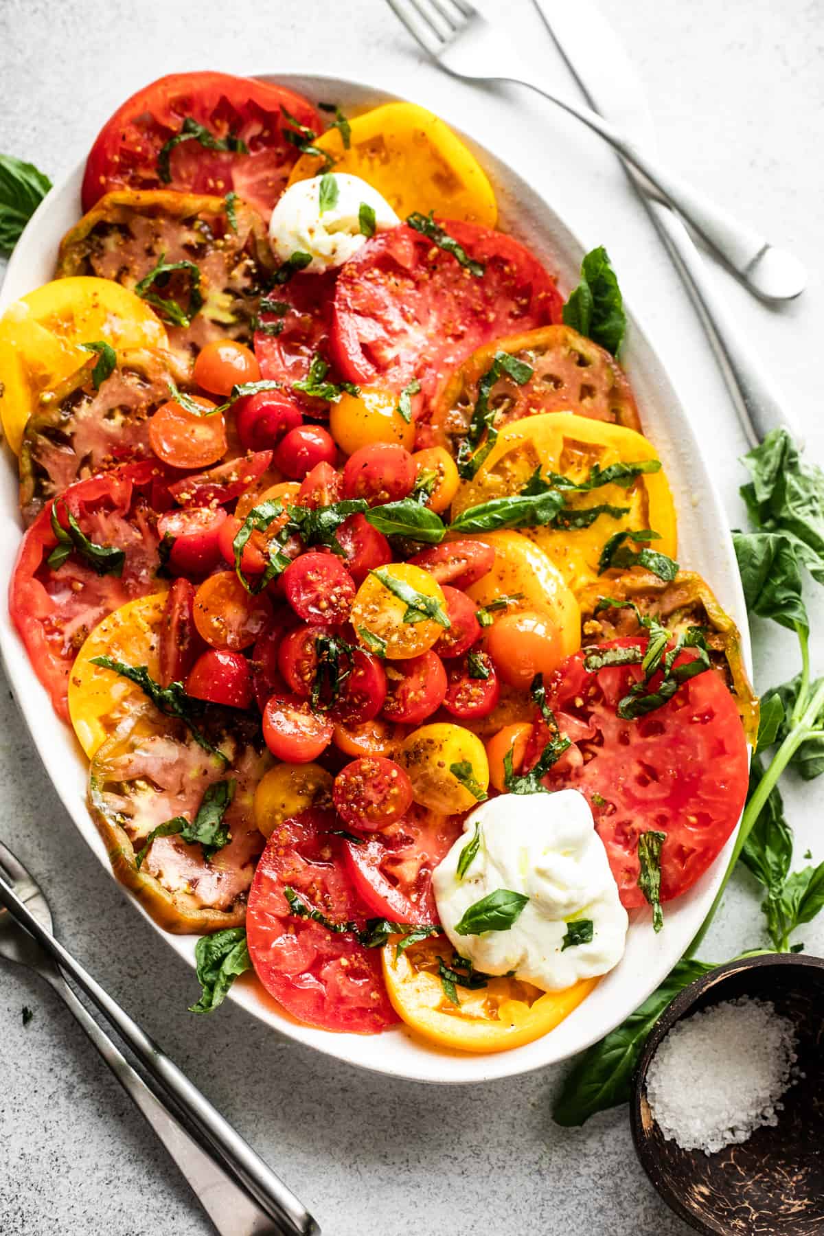 slices of heirloom tomatoes arranged on an oval platter, with halved cherry tomatoes placed in the center, burrata cheese at the top of the plate and the bottom, and a garnish of basil over the entire salad.