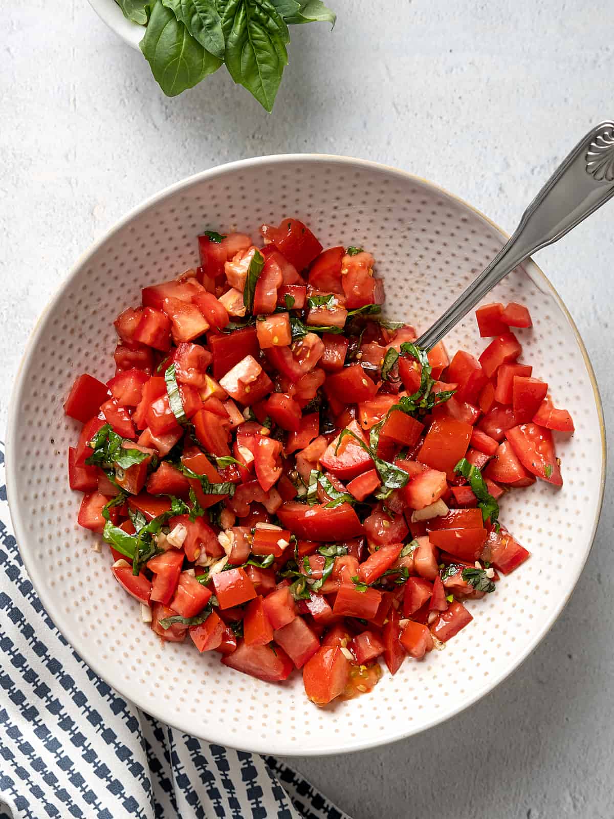A mixture of diced tomato, chopped basil and minced garlic in a bowl with a spoon.
