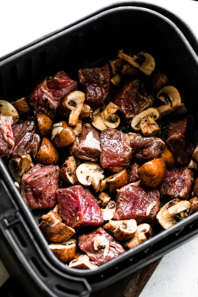 raw steak chunks and quartered bella mushrooms in a black air fryer basket.
