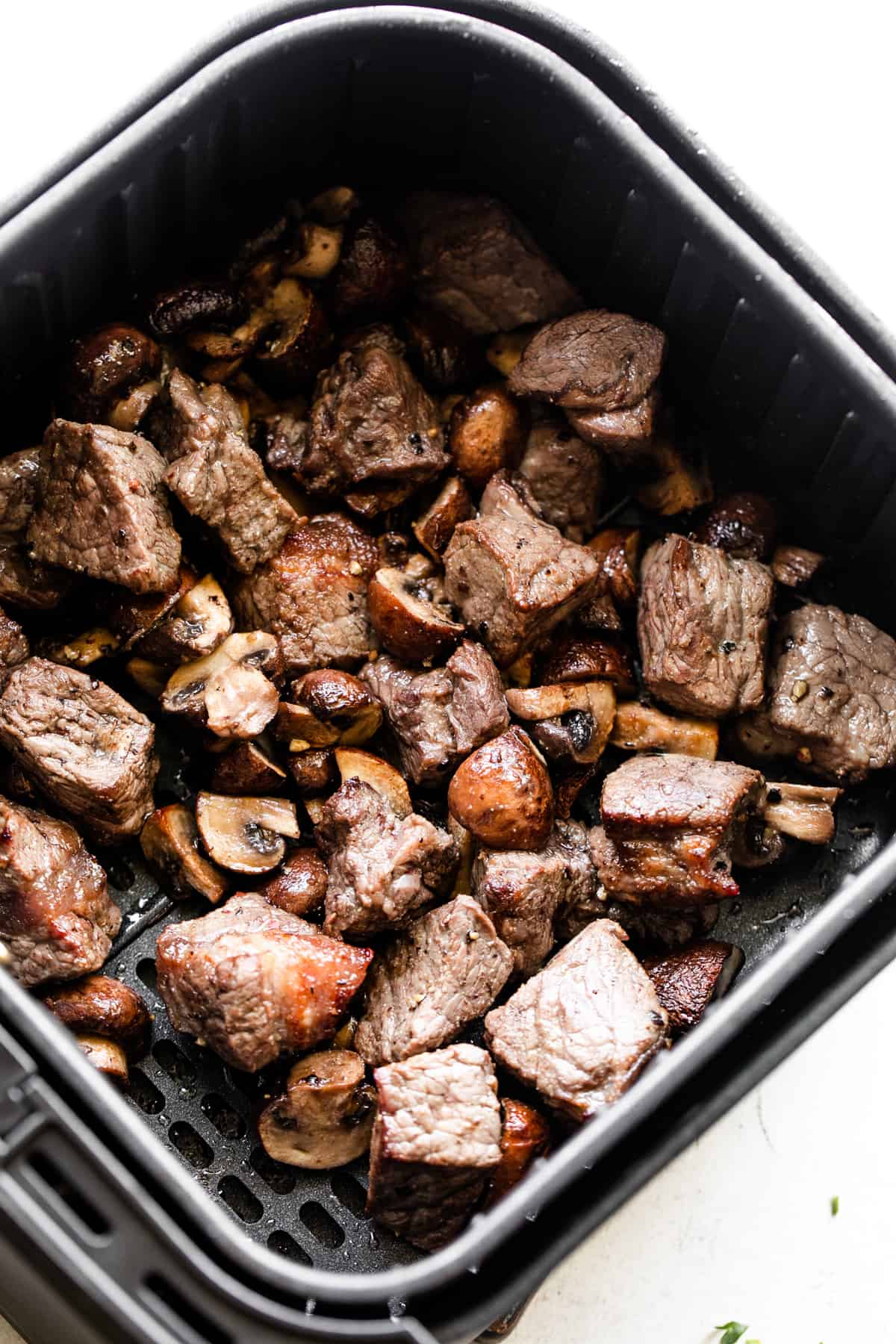 overhead shot of cooked steak bites and quartered bella mushrooms in a black air fryer basket.
