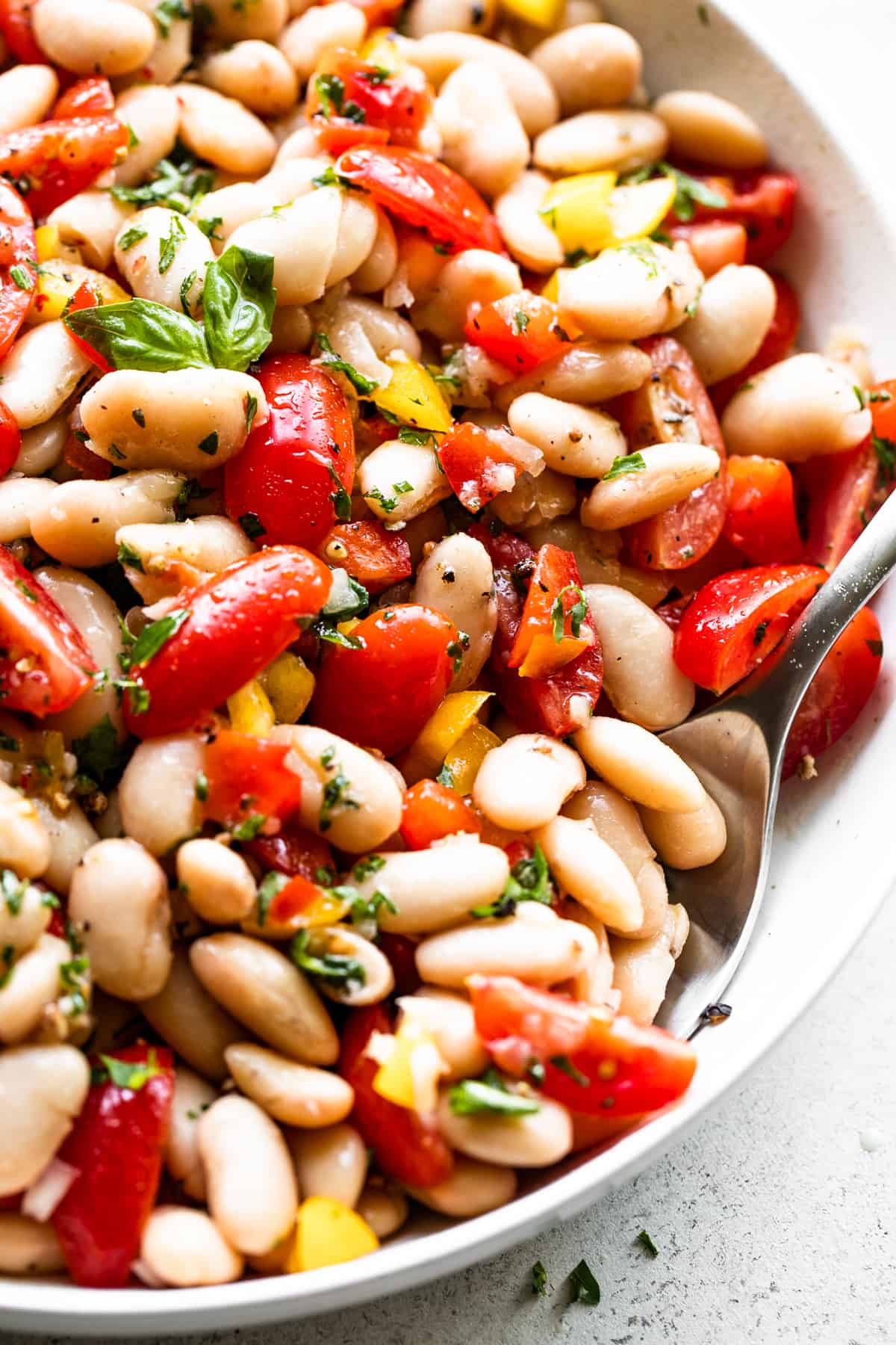 close up overhead shot of a salad bowl with cannellini beans, tomatoes, bell peppers, and a spoon set inside the right bottom side of the bowl.