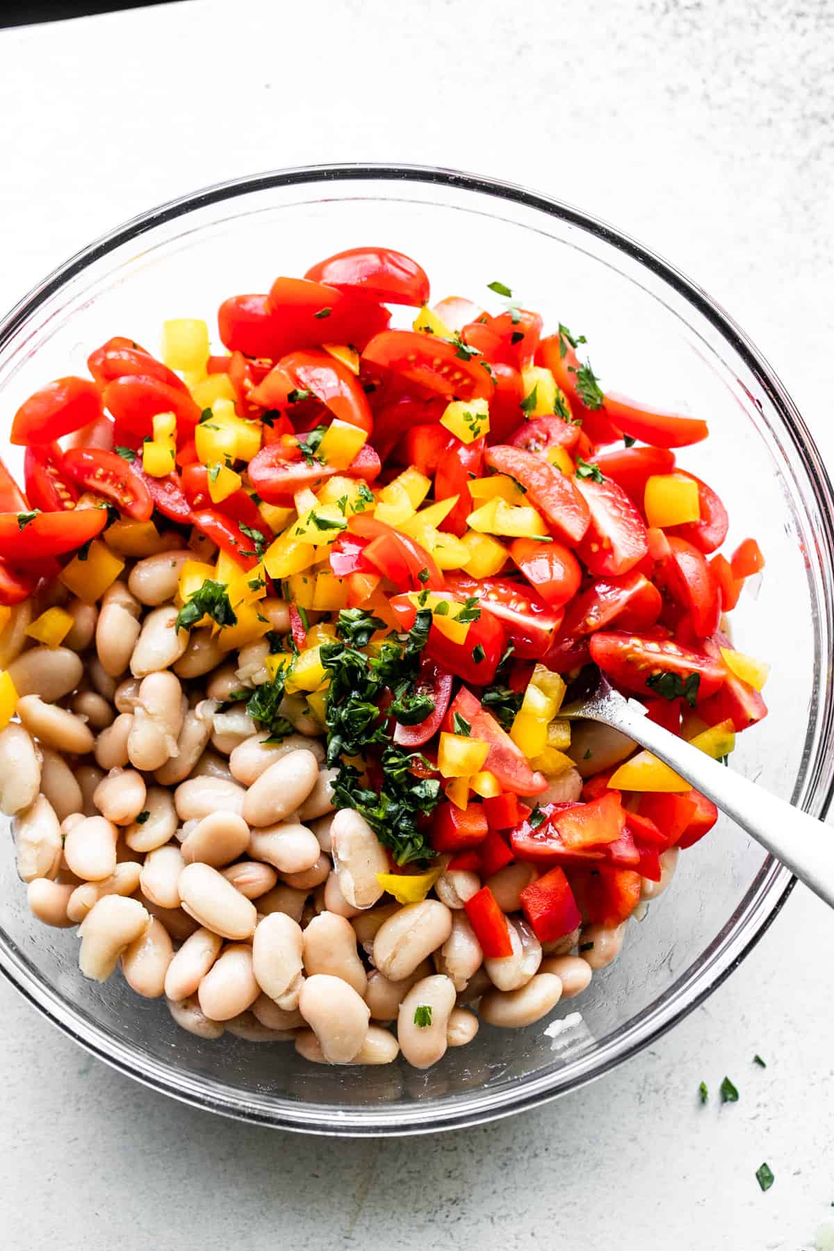 overhead shot of a large glass bowl filled with cannellini beans, halved red cherry tomatoes, diced red bell pepper, diced yellow bell pepper, and a sprinkle of chopped fresh basil set in the center.
