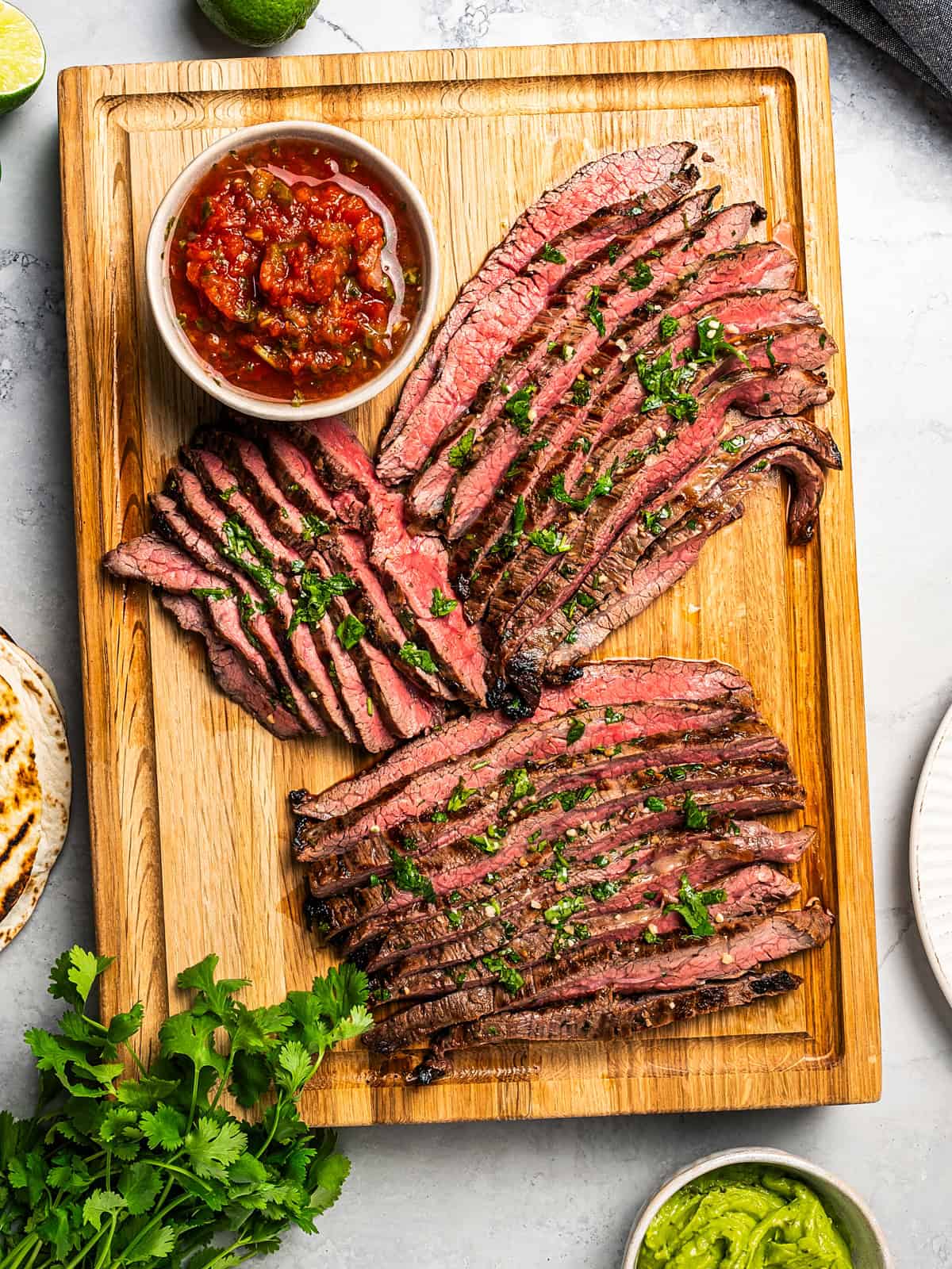 Slices of Carne Asada on a wooden cutting board. A bowl of pico de gallo is also set near the meat.