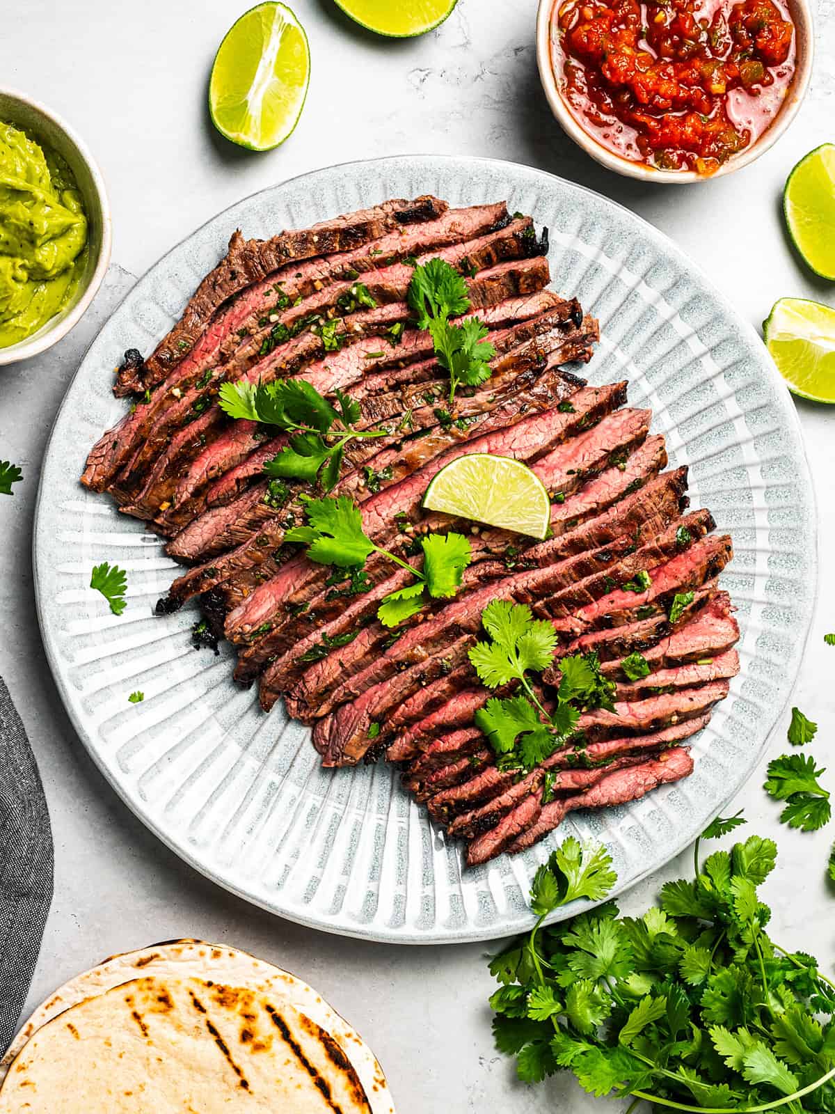 Grilled carne asada cut into thin slices and served on a platter with a garnish of cilantro and lime wedges.