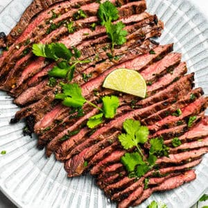 Grilled flank steak cut into thin slices and served on a platter with a garnish of cilantro and lime wedges.