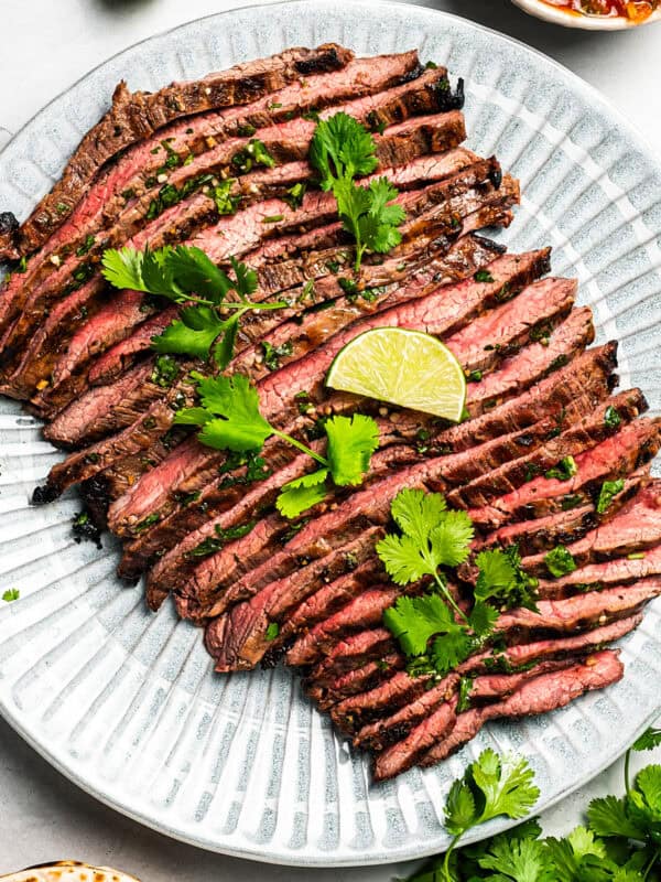 Grilled flank steak cut into thin slices and served on a platter with a garnish of cilantro and lime wedges.