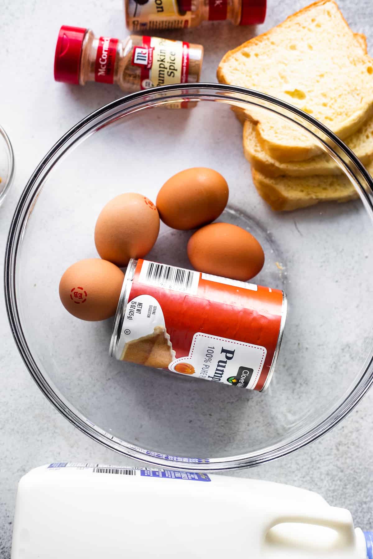 four eggs and can of pumpkin puree inside of a glass mixing bowl, and slices of bread and a cinnamon shaker placed right above the mixing bowl.