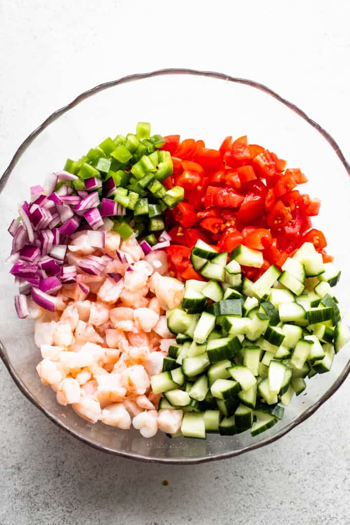 a glass mixing bowl with diced cucumbers, diced tomatoes, diced avocados, diced shrimp, diced red onions, and diced jalapenos marinated in citrus juice.