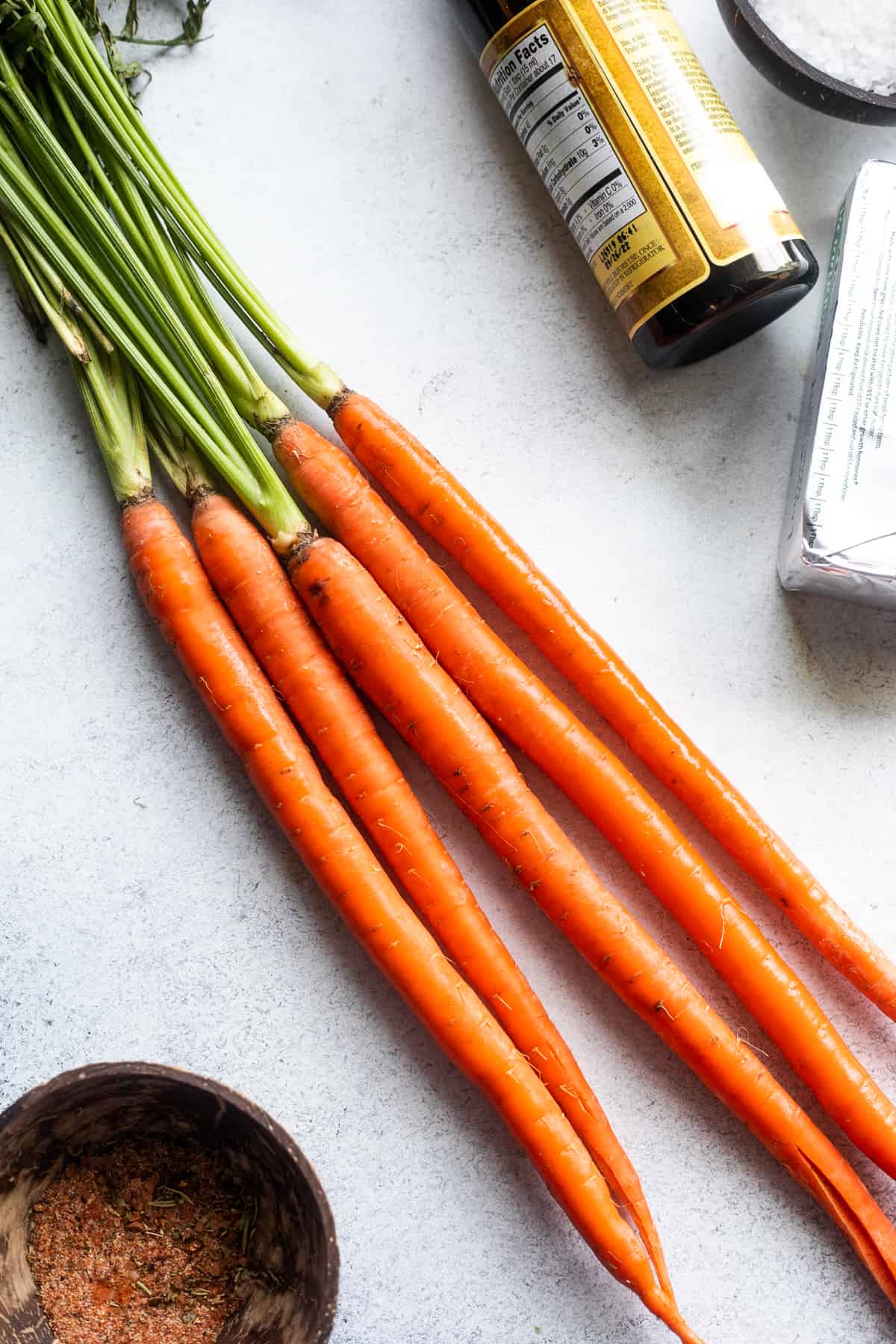 Give long carrots with green stalks arranged on a white background with balsamic vinegar and a seasonings mixture placed around the carrots.