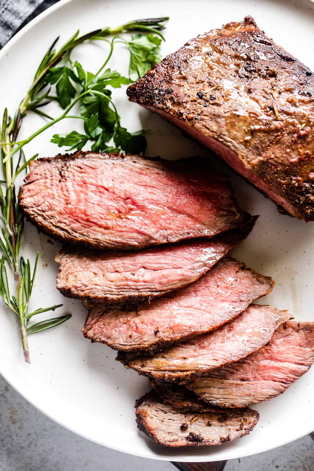 overhead shot of partially sliced sirloin tip roast on a white plate next to rosemary sprigs.