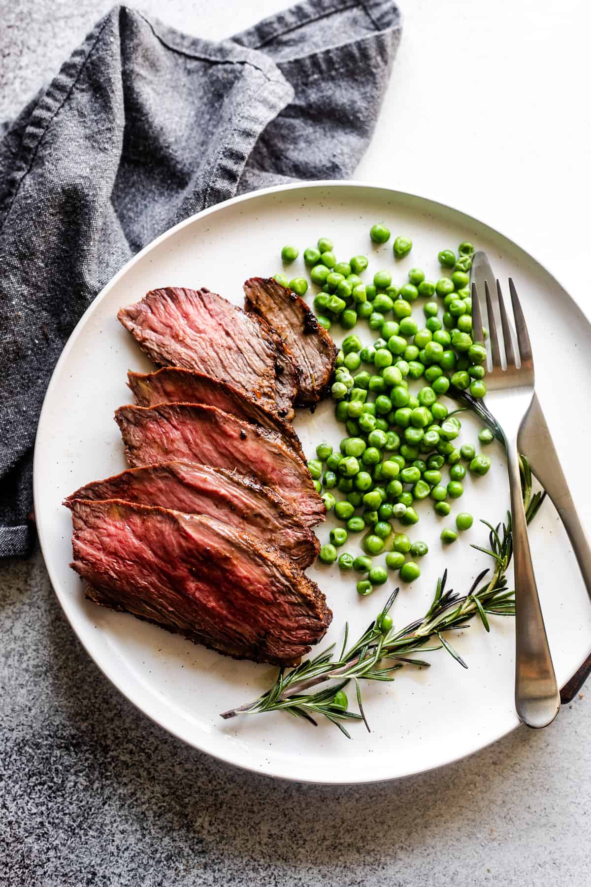 slices of sirloin tip roast arranged on a plate with green peas and a rosemary branch served next to it.