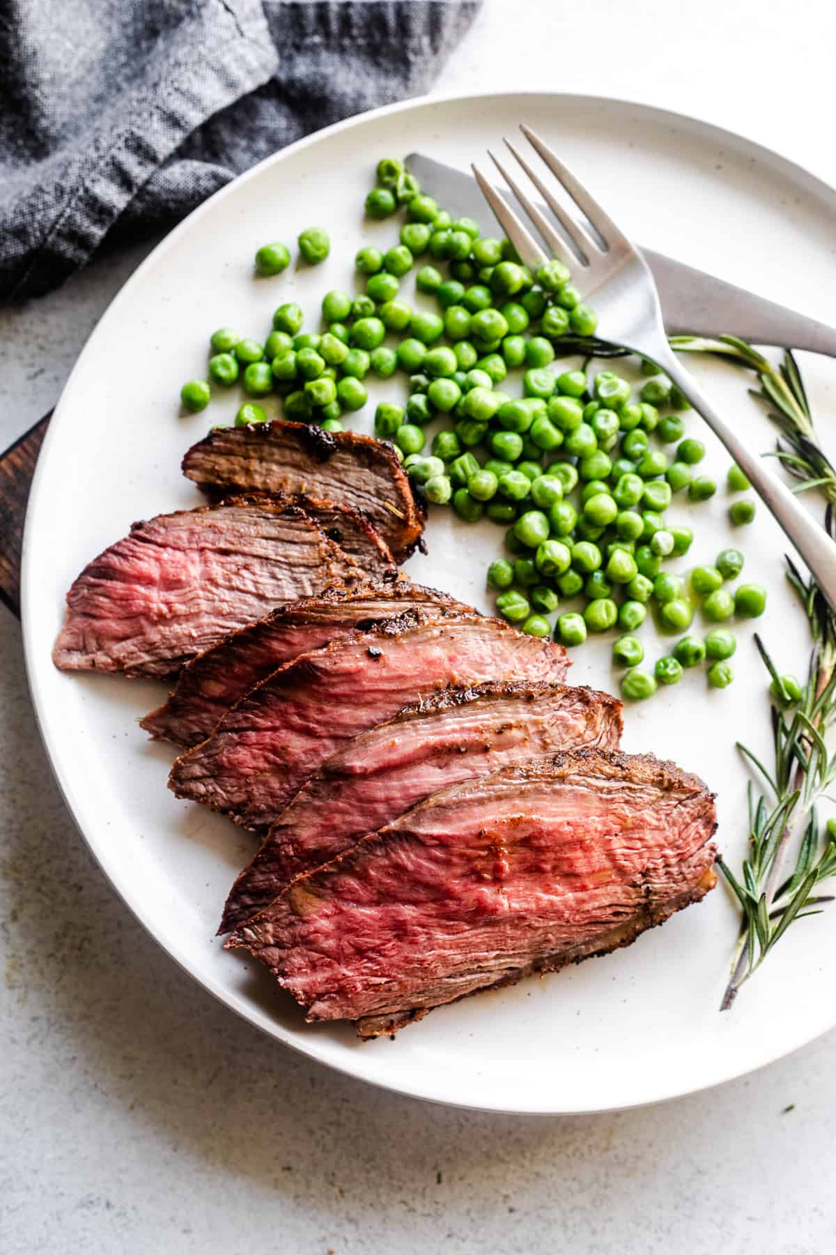 slices of sirloin tip roast arranged on a plate with green peas and a rosemary branch served next to it.