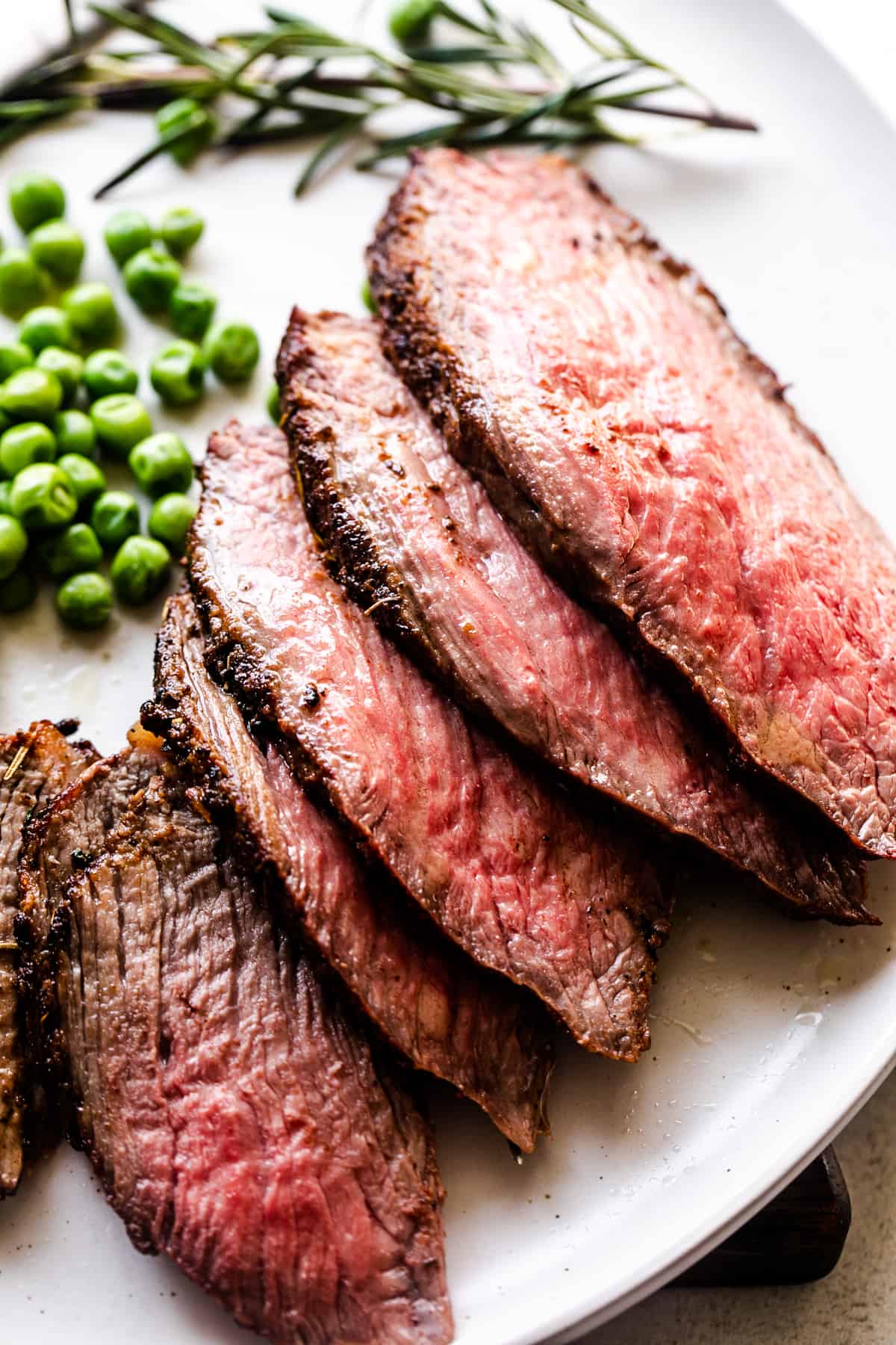 five thinly sliced pieces of tri tip arranged on a white plate with green peas next to it and a branch of rosemary.