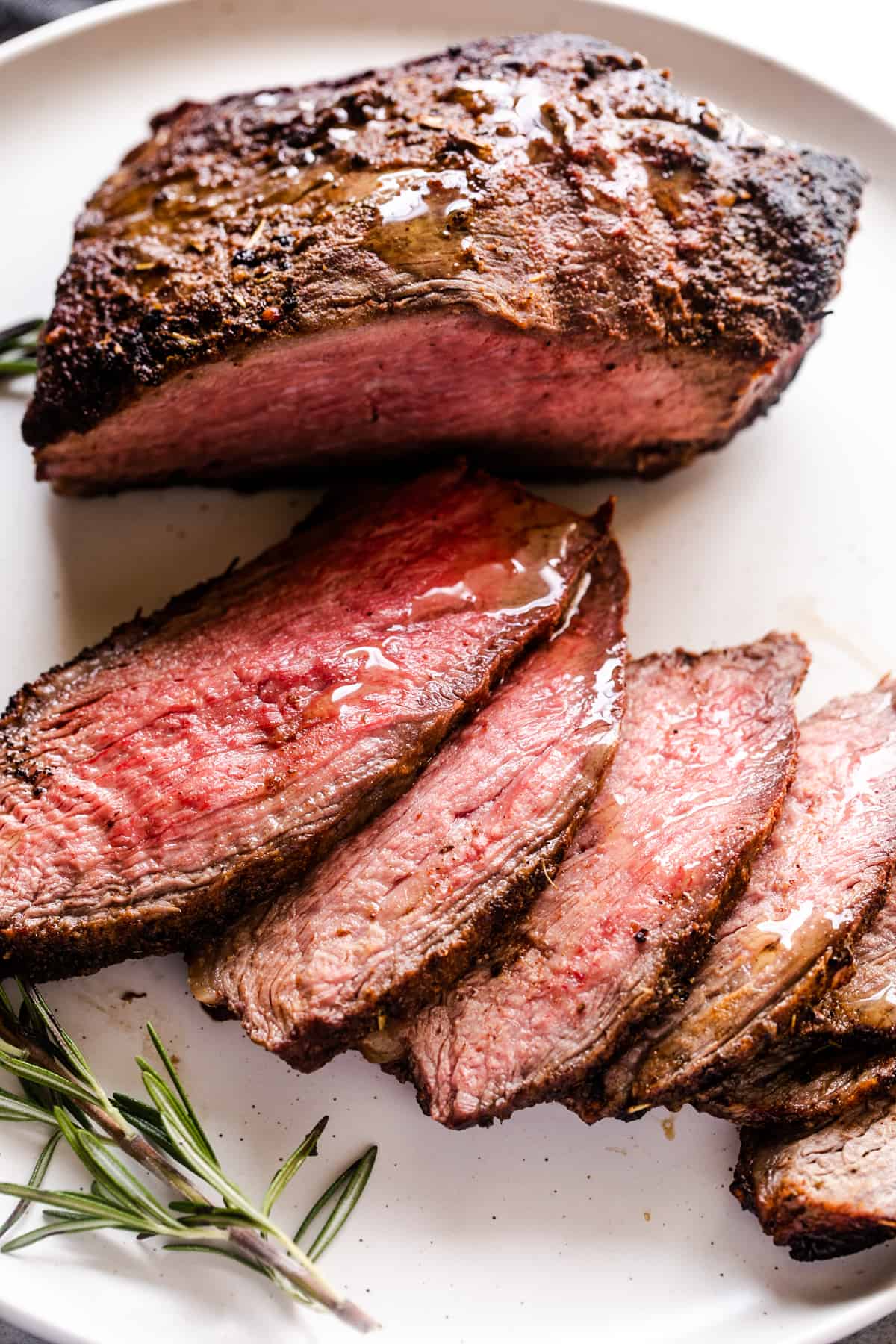 overhead shot of partially sliced sirloin tip roast on a white plate next to a rosemary sprig.