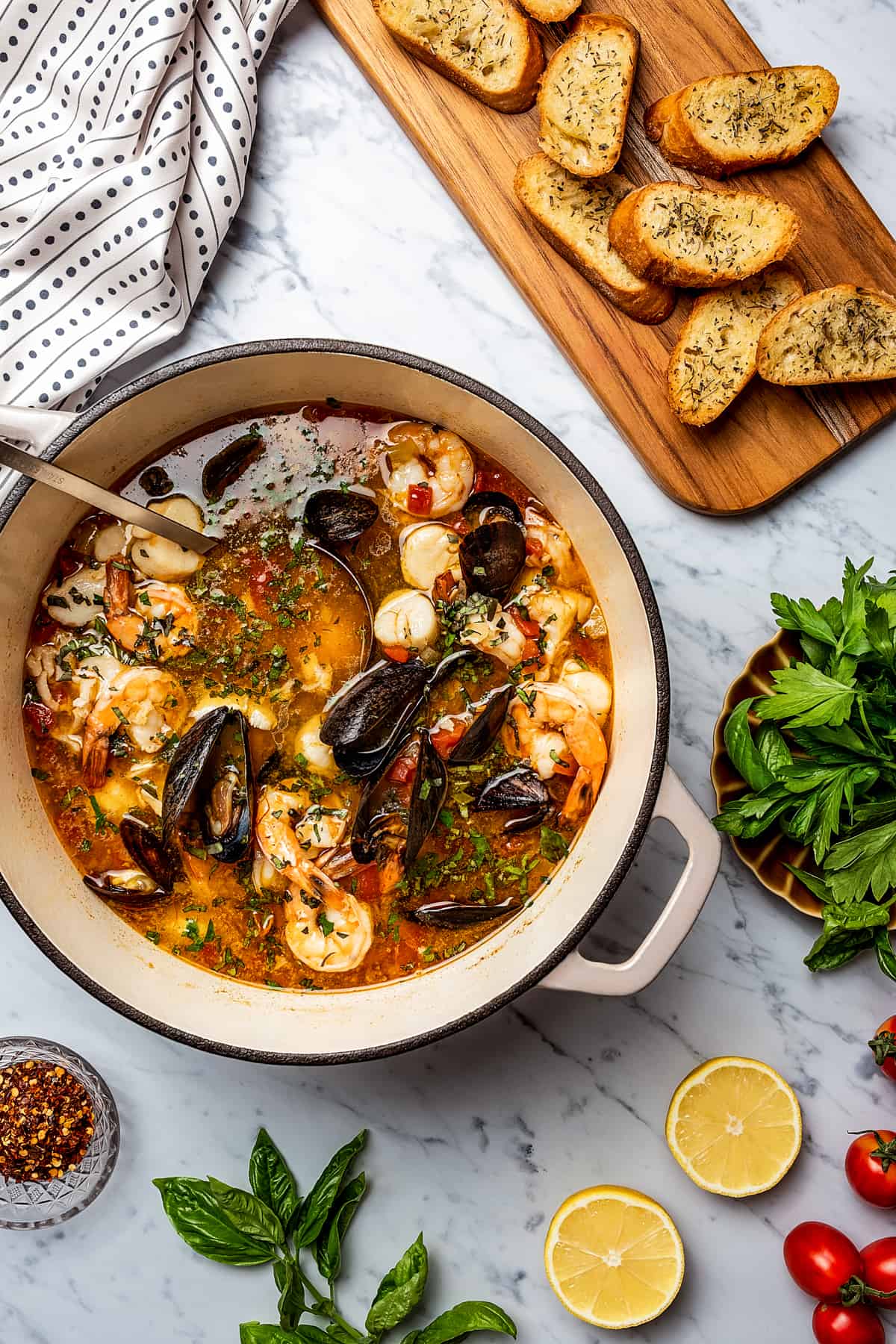 Overhead shot of seafood stew in a pot, next to a cutting board of toasted baguette slices. Various other ingredients are arranged on the work surface.