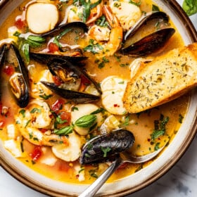 Bouillabaisse in a soup bowl, with a spoon and slice of toasted baguette.