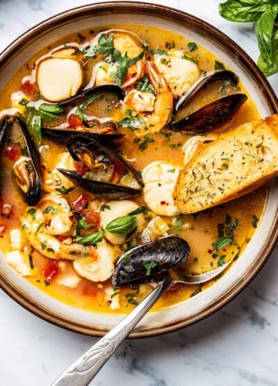 Bouillabaisse in a soup bowl, with a spoon and slice of toasted baguette.