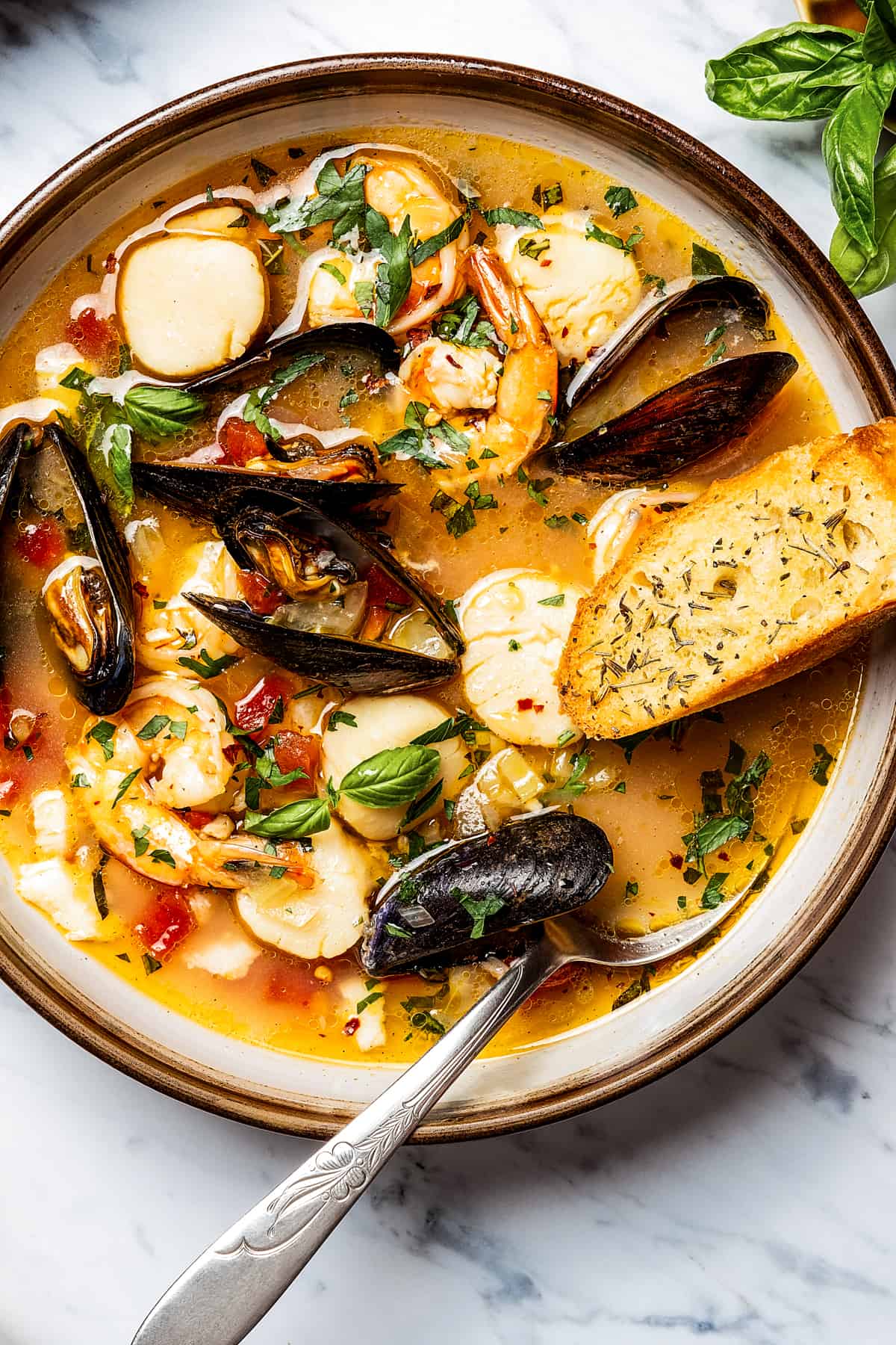 Bouillabaisse in a soup bowl, with a spoon and slice of toasted baguette.