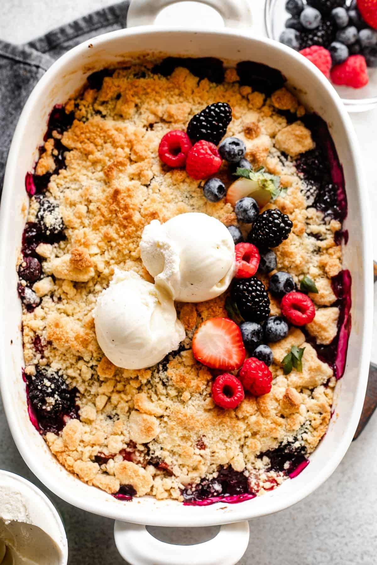 overhead shot of a berry cobbler topped with a crumbly biscuit topping, a row of fresh berries, and two scoops of vanilla ice cream.