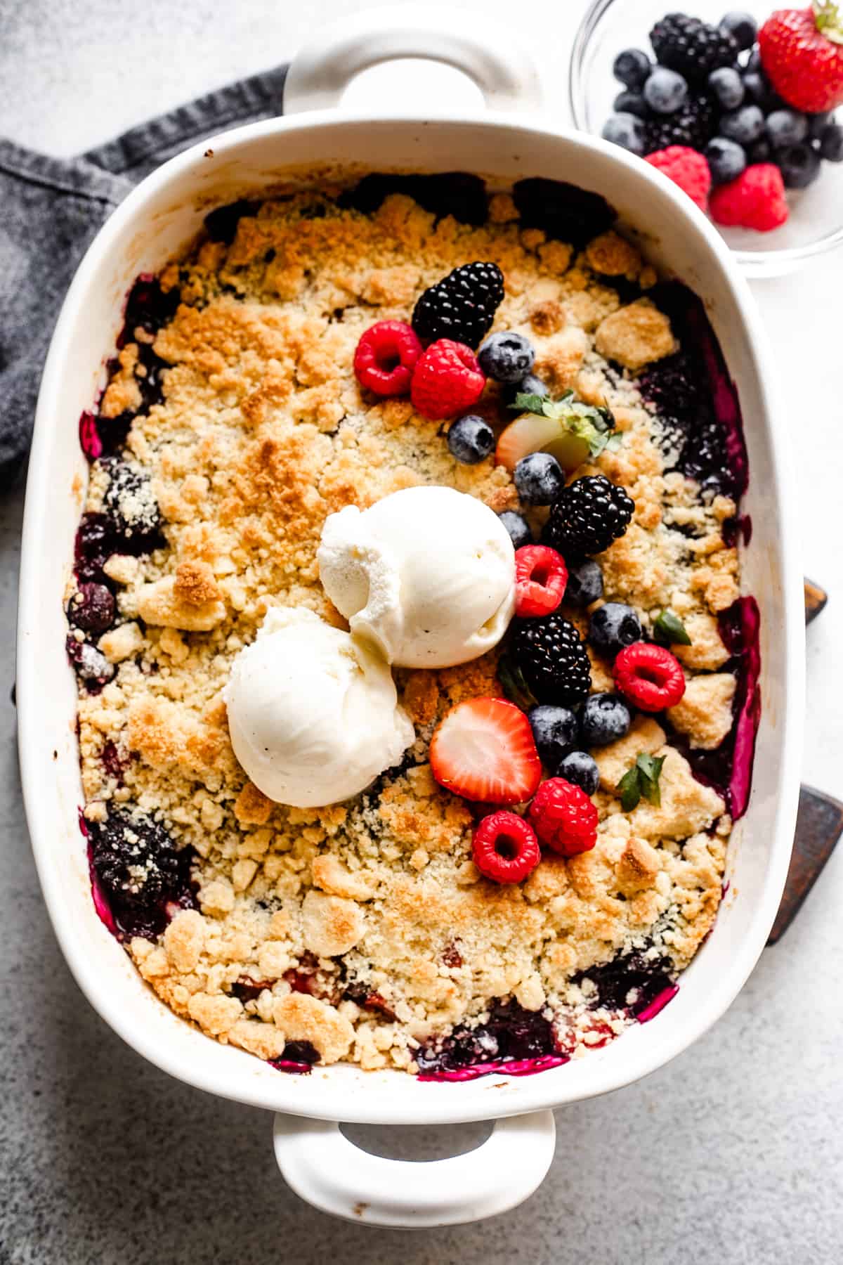 overhead shot of a berry cobbler topped with a crumbly biscuit topping, a row of fresh berries, and two scoops of vanilla ice cream.