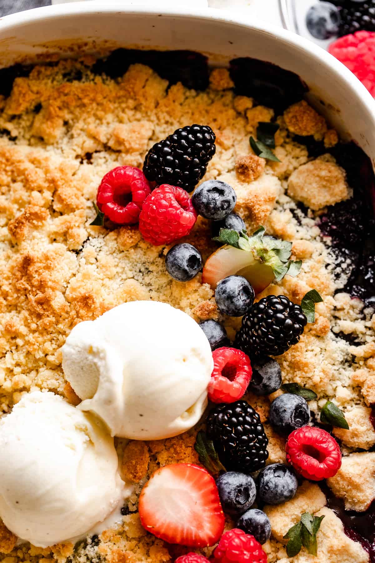 close up shot of fresh berries and two ice cream scoops arranged on top of a crumbly biscuit topping.