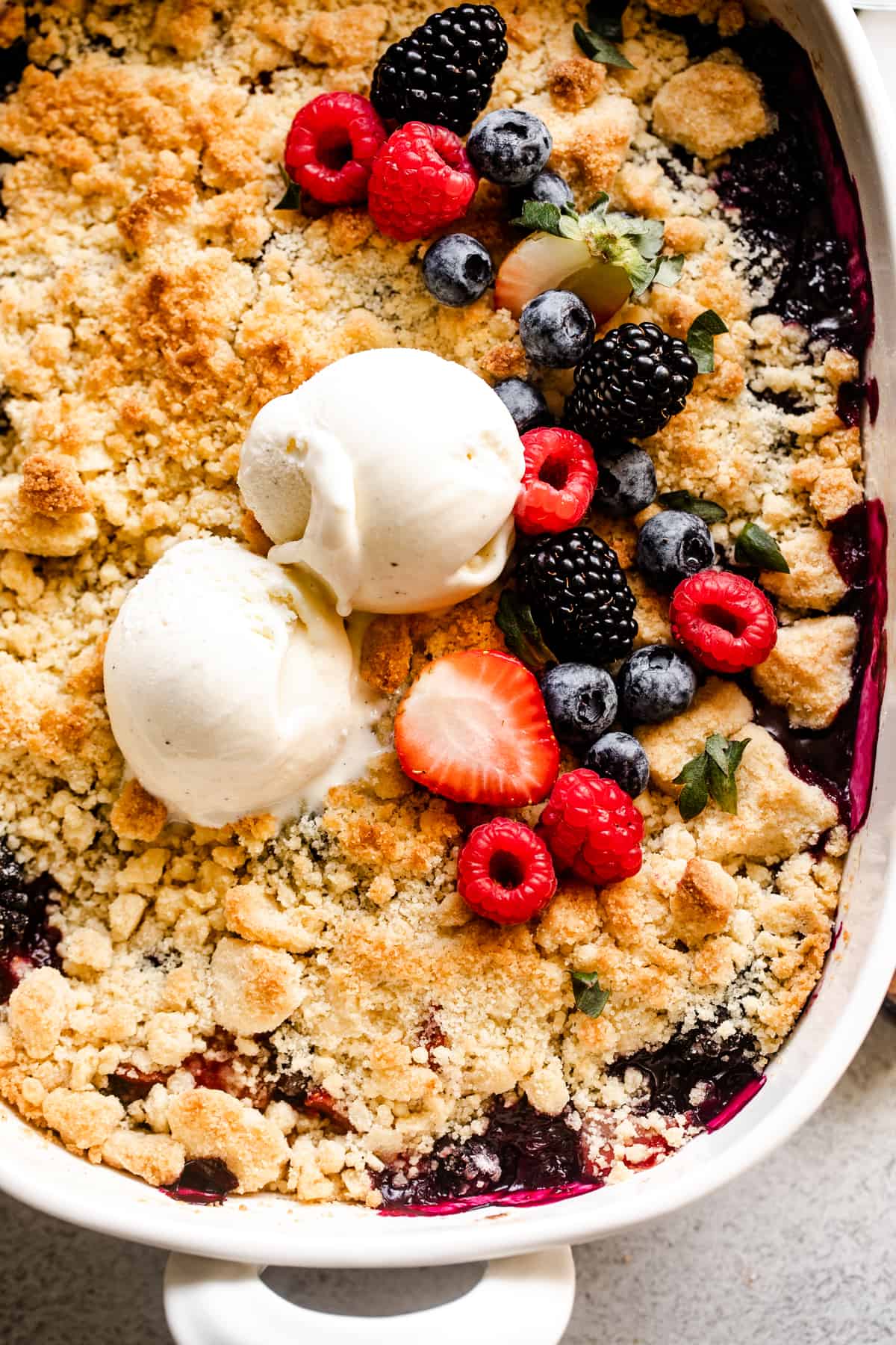 overhead shot of a berry cobbler topped with a crumbly biscuit topping, a row of fresh berries, and two scoops of vanilla ice cream.