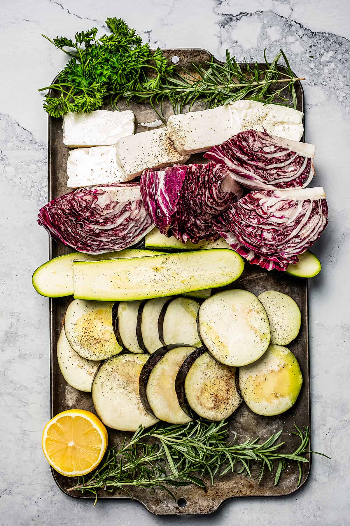 A sheet pan with layers of herbs, sliced cheese, vegetables, and a lemon slice.
