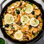 A skillet of pasta and shrimp, with lemon halves, herbs, herbs, and feta arranged on the table.