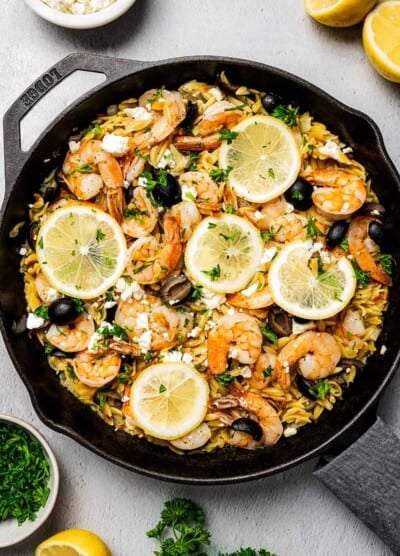 A skillet of pasta and shrimp, with lemon halves, herbs, herbs, and feta arranged on the table.