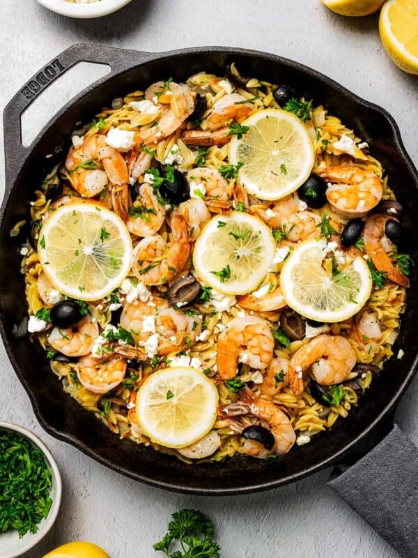 A skillet of pasta and shrimp, with lemon halves, herbs, herbs, and feta arranged on the table.