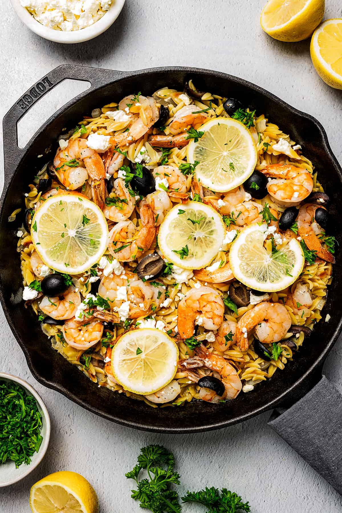 A skillet of pasta and shrimp, with lemon halves, herbs, herbs, and feta arranged on the table.