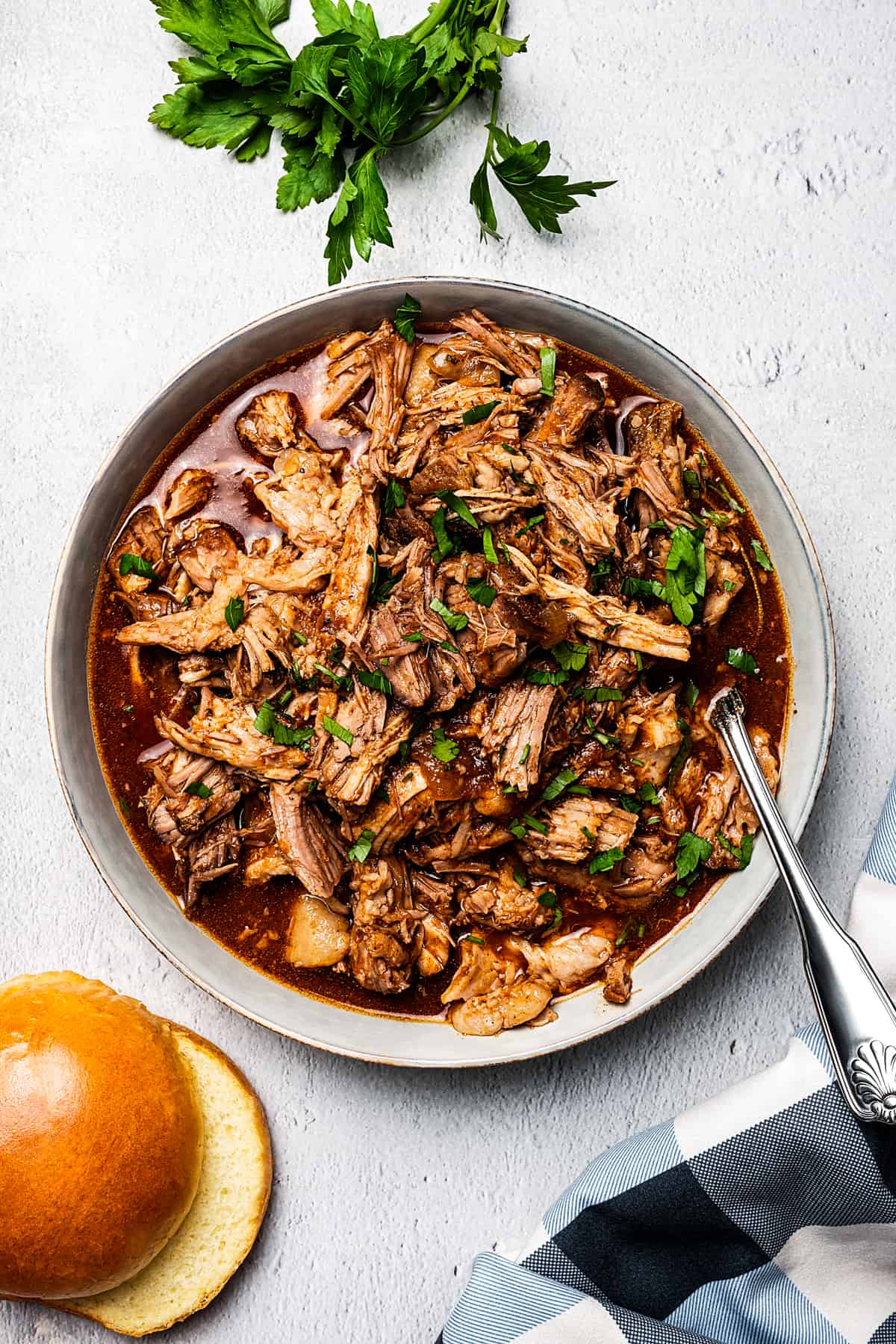 overhead shot of slow cooked pulled pork in a bowl, with burger buns placed to the bottom left of the bowl.