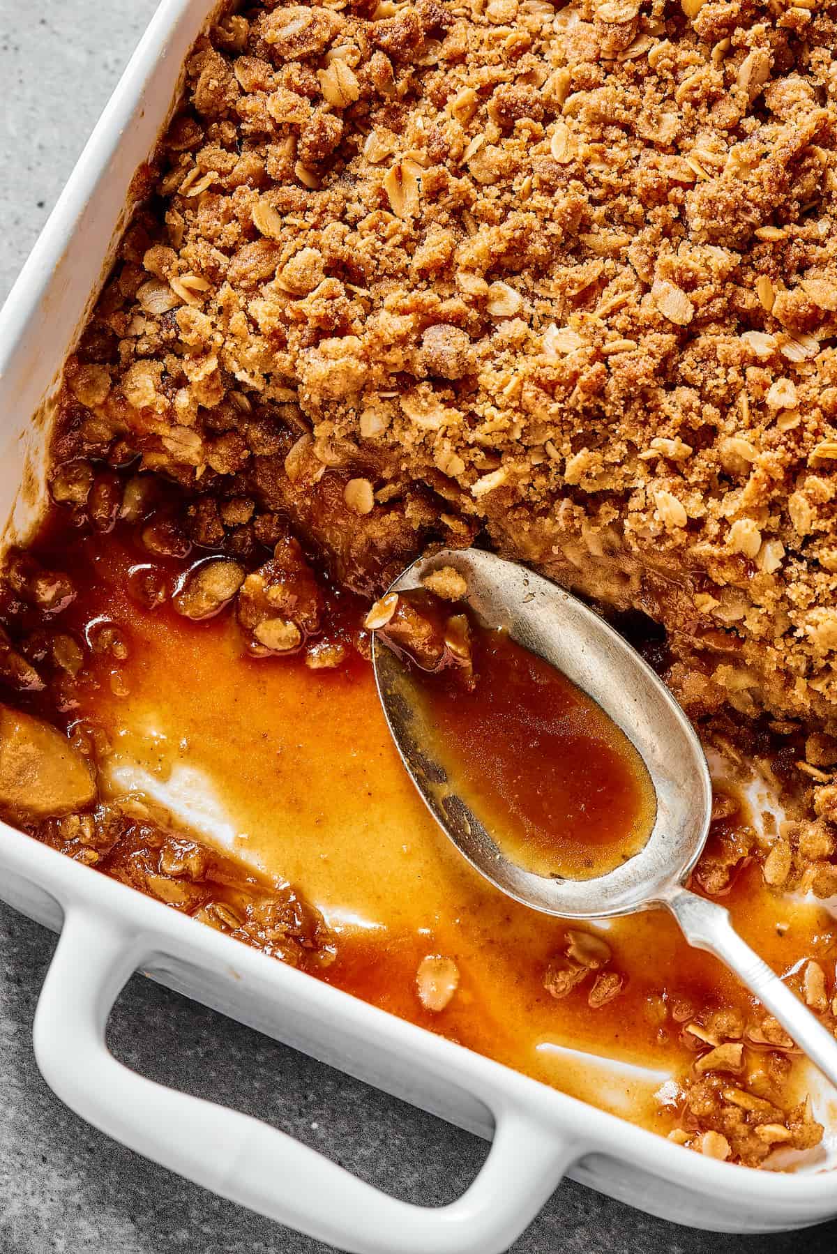 Close-up shot of the corner of a white casserole dish with an apple dessert, showing the apples, topping, and syrup. A spoon is resting in the empty part of the casserole dish.