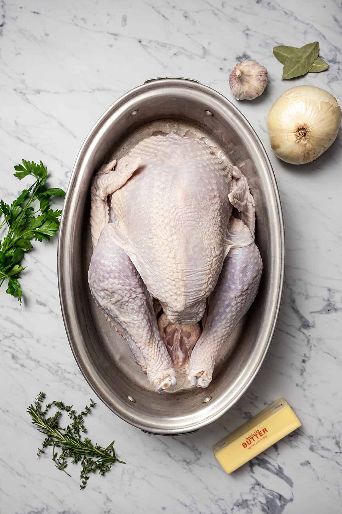 A whole turkey in a roasting pan. Fresh herbs, garlic, and butter are on the work surface.