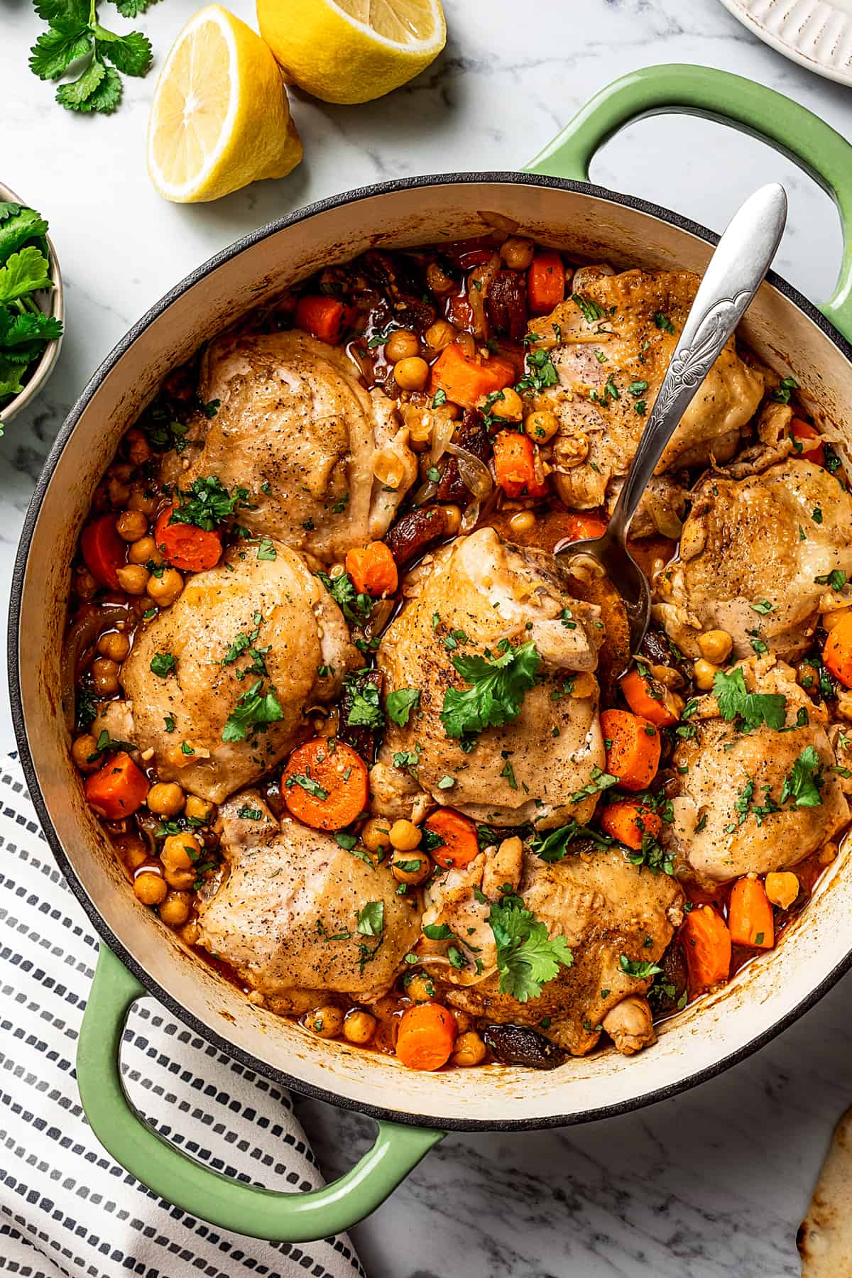 A skillet of chicken thighs in sauce, with fresh herbs and lemon on the work surface.