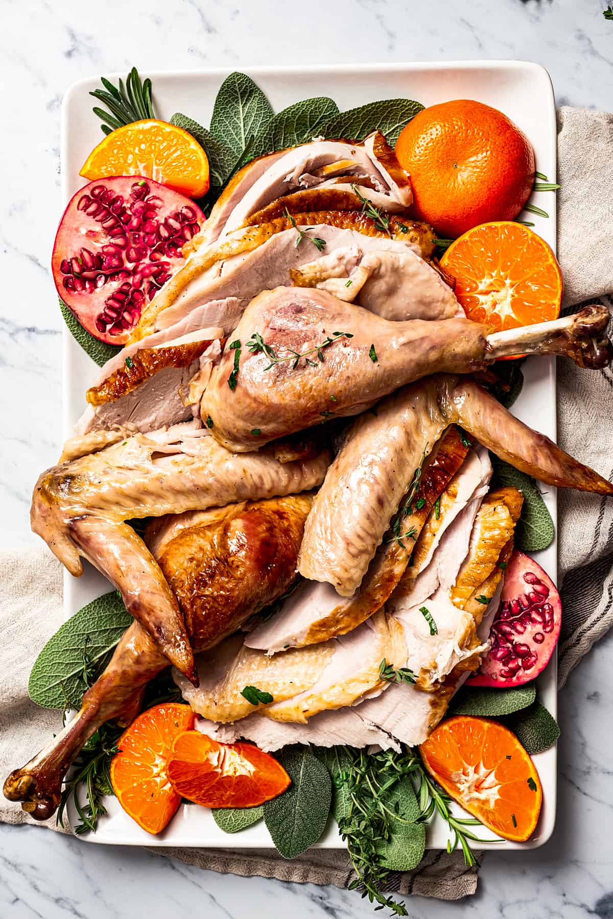 A serving platter of turkey drumsticks, wings, and breast meat, garnished with pomegranate and orange sections and fresh herbs.