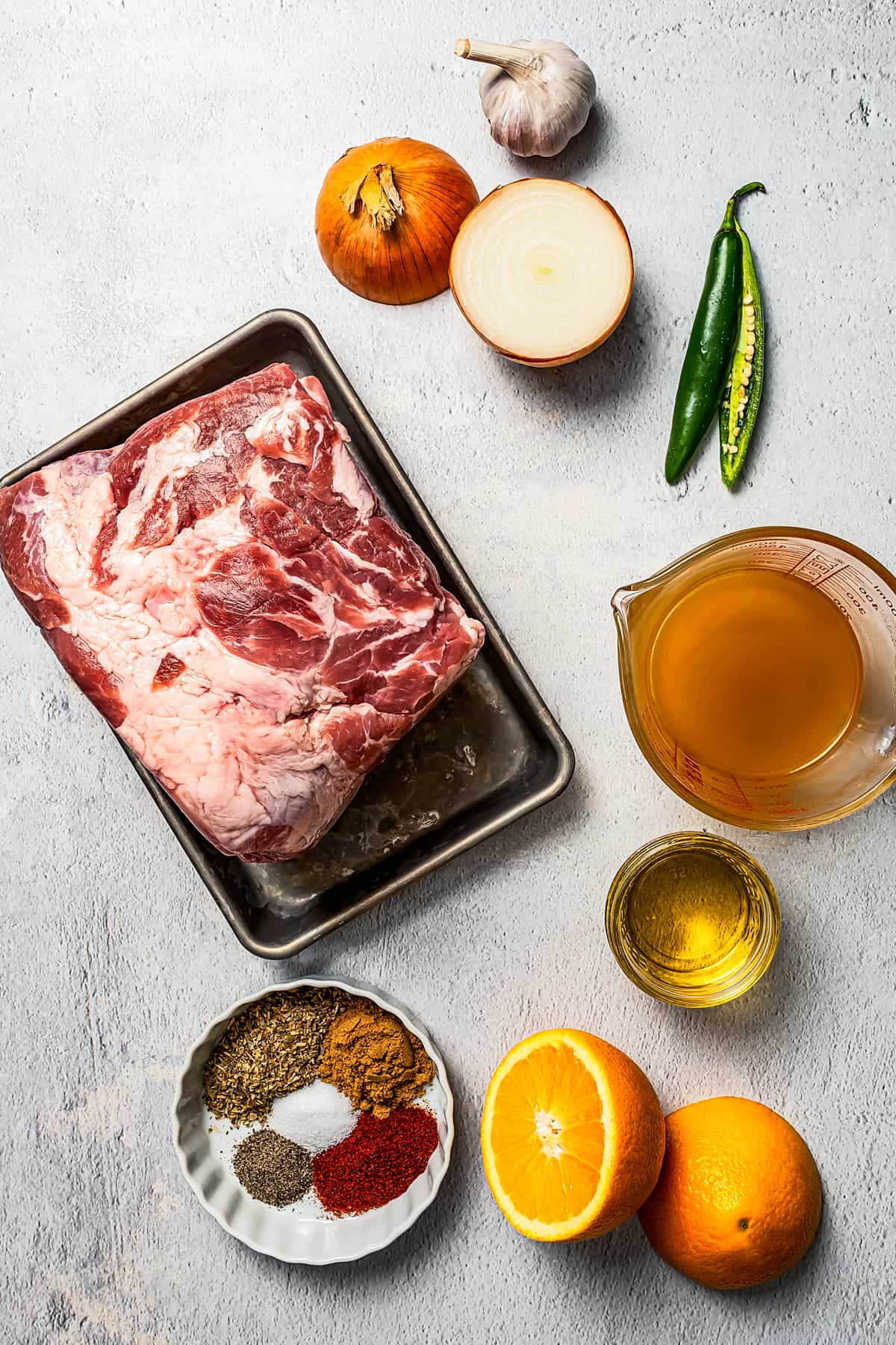 Ingredients for Pernil, pictured from top: Garlic, onion, chili peppers, pork shoulder, broth, seasonings, oil, oranges.
