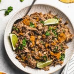 overhead shot of shredded pork pernil in a white serving dish, and garnished with lime slices plus parsley. Corn tortillas are placed to the right, near the serving dish, and a bowl of rice is placed on the opposite side.