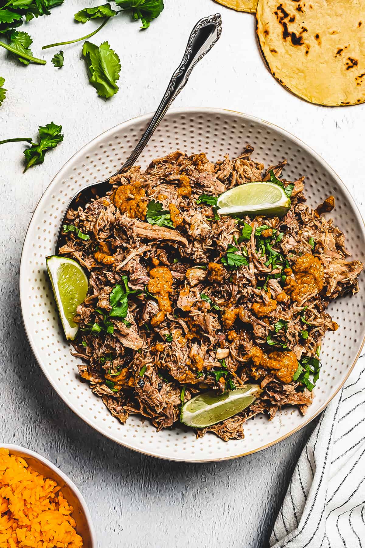 overhead shot of shredded pork pernil in a white serving dish, and garnished with lime slices plus parsley. Flour tortillas are placed to the right, near the serving dish, and a bowl of rice is placed on the opposite side.