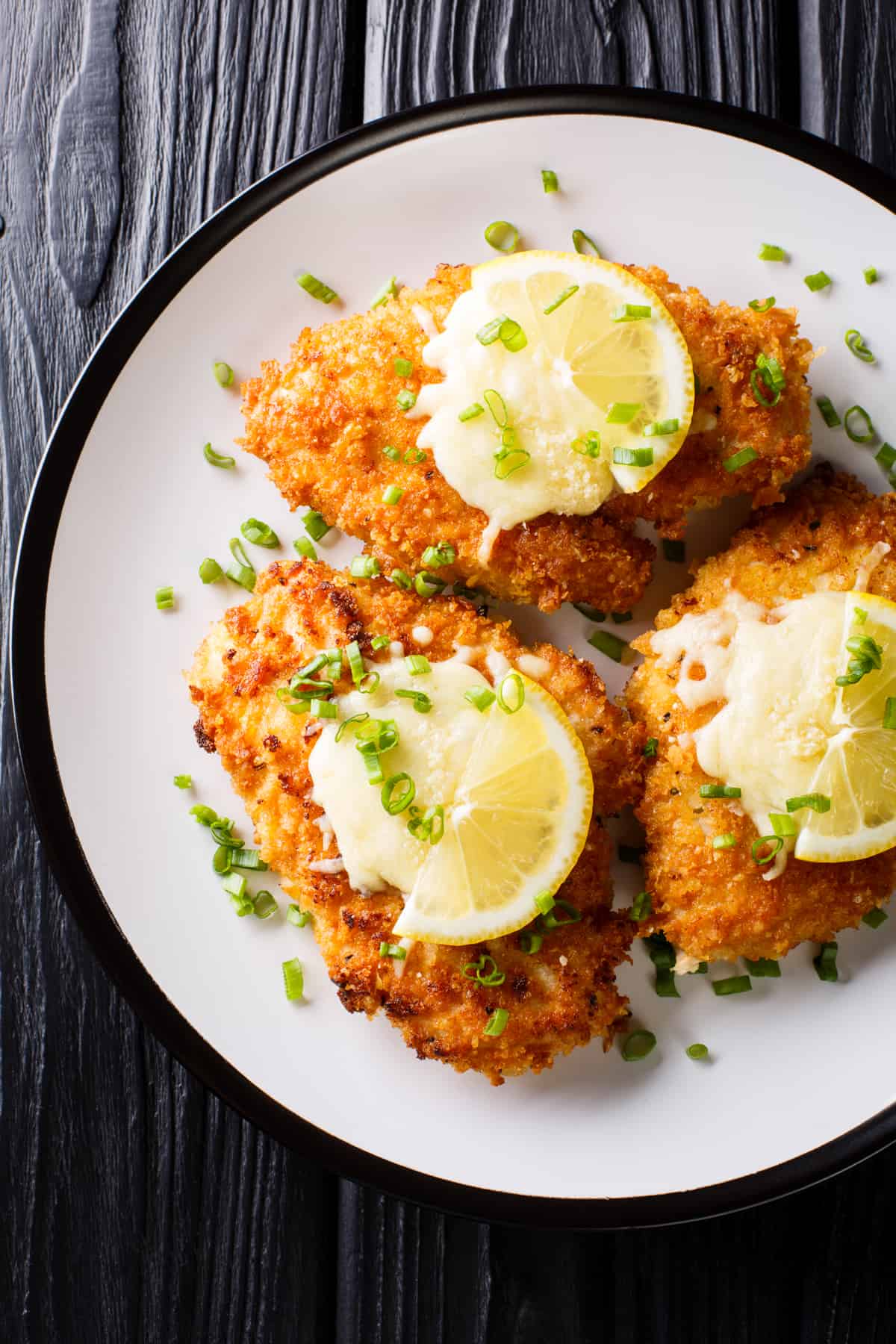 overhead shot of three breaded chicken breasts, also known as chicken milanese, arranged on a plate and topped with lemon slices.