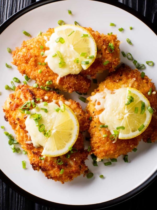 overhead shot of three breaded chicken breasts, also known as chicken milanese, arranged on a plate and topped with lemon slices.