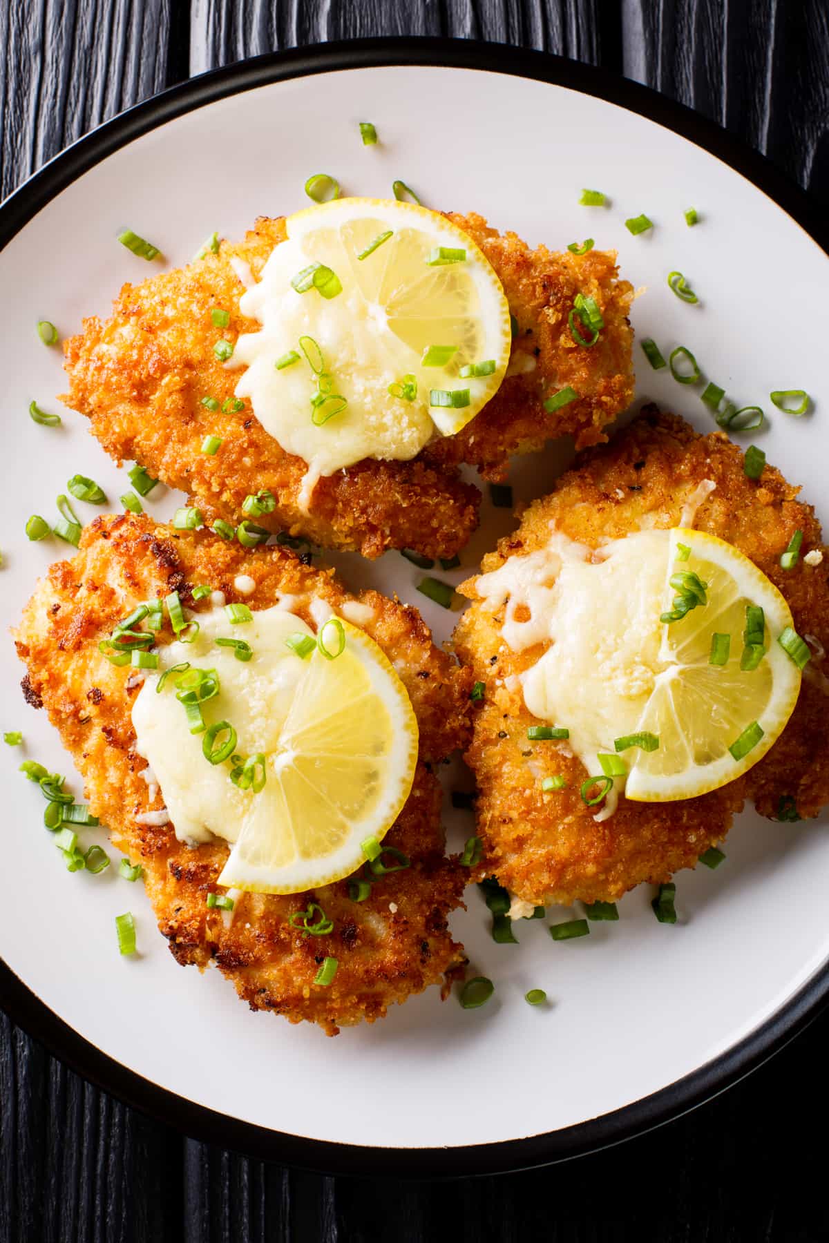 Overhead shot of three breaded chicken breasts, also known as chicken milanese, arranged on a plate and topped with lemon slices and chives.
