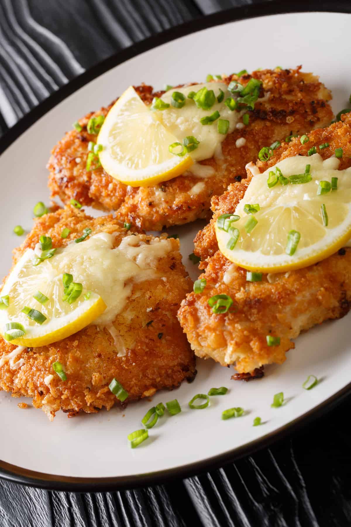 three breaded chicken breasts arranged on a plate and topped with lemon slices.