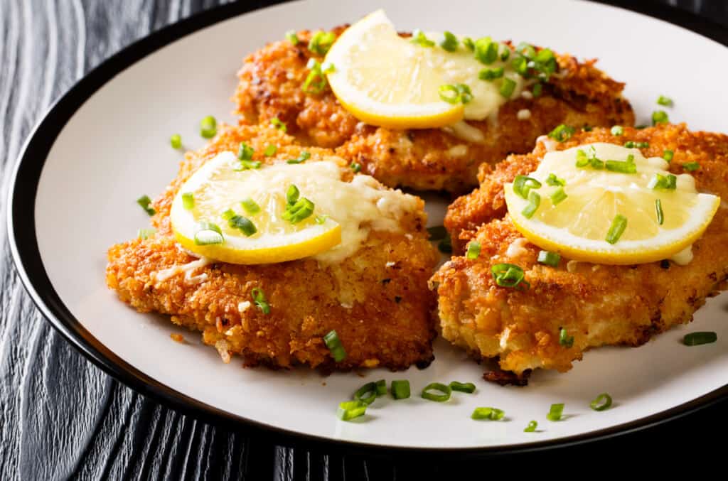 three breaded chicken breasts arranged on a plate and topped with lemon slices.