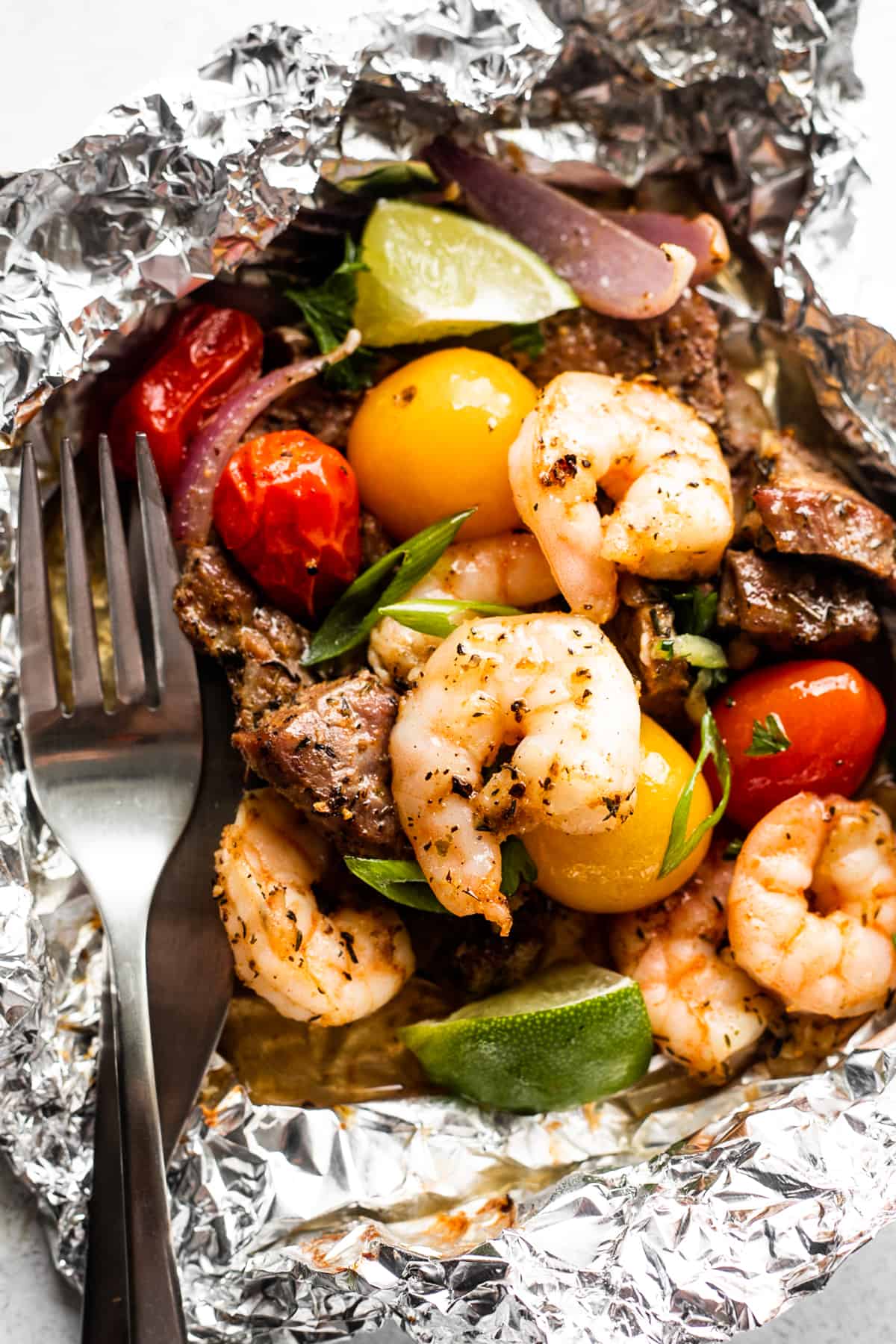 close up shot of a foil pack filled with steak bites, shrimp, red onions, and colored grape tomatoes, and a fork set on the left side of the packet.