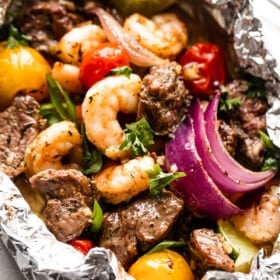 close up shot of a foil pack filled with steak bites, shrimp, red onions, and colored grape tomatoes.